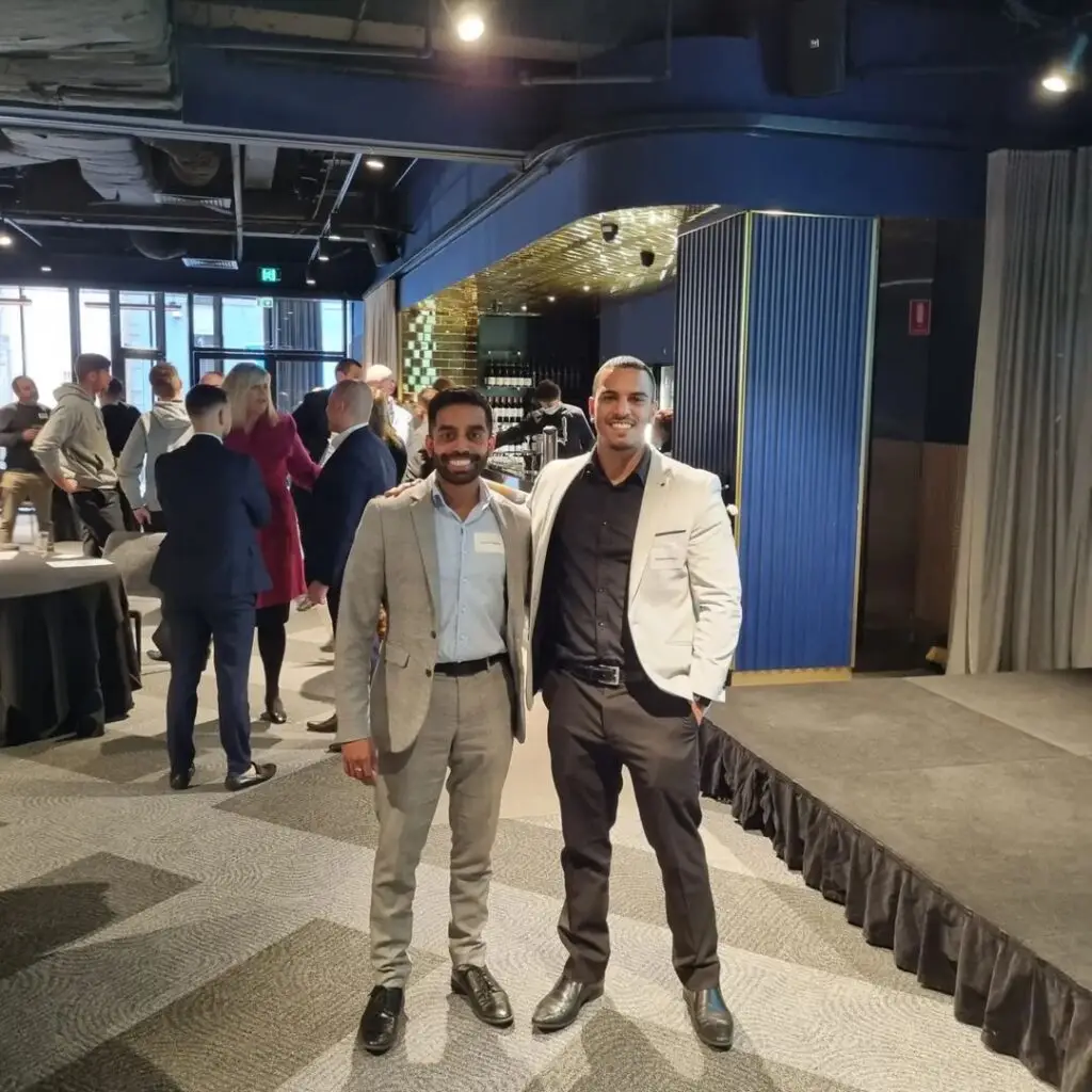 Two men standing together in a business event setting, with a testimonial from Angelo Perera & Trevor Anthony, Directors of Situate Finance & Carvera Group, commending the quality and organization of video content produced weekly.