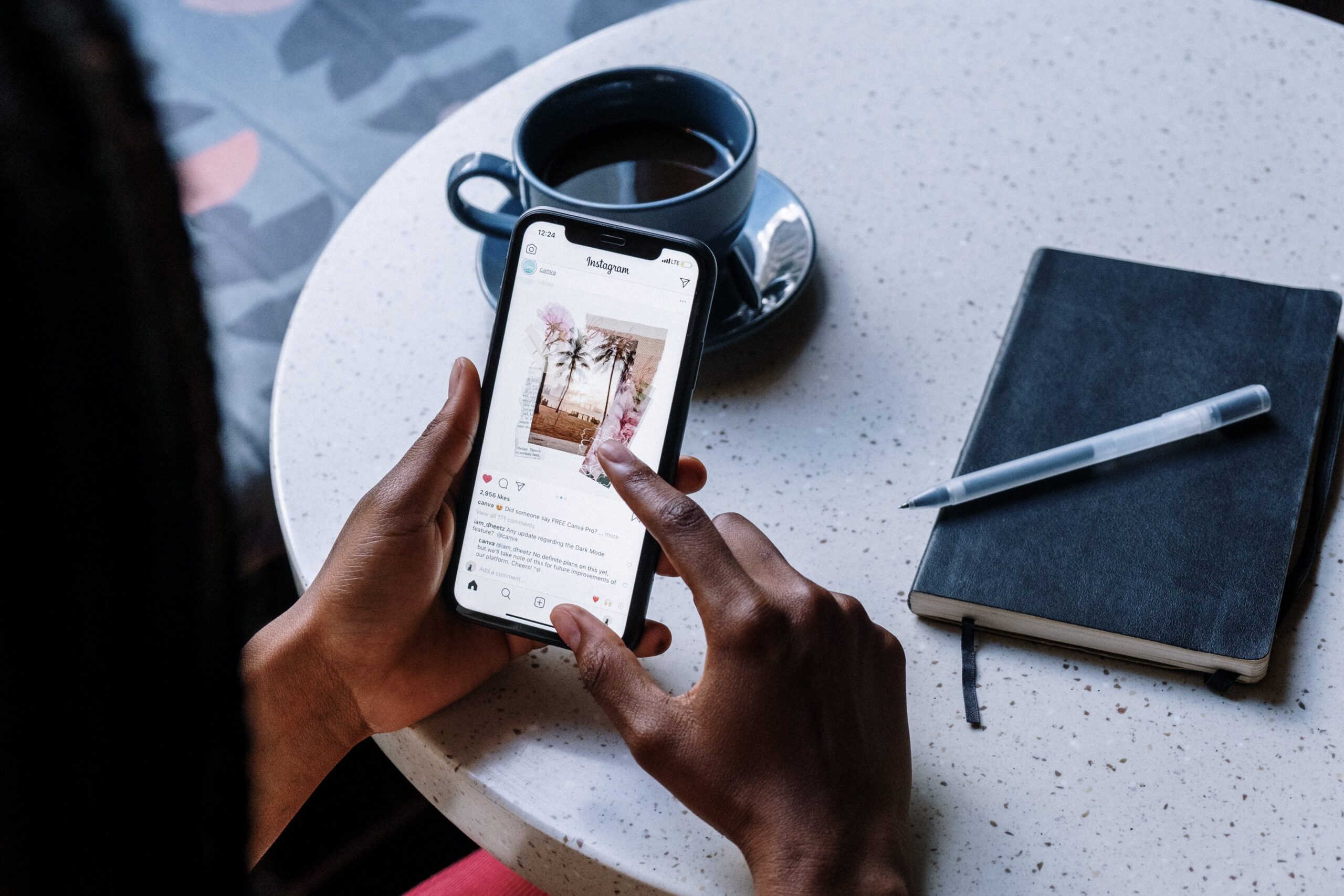 A person's hands holding a smartphone displaying an Instagram post, with the phone resting on a table next to a cup of coffee and an open notebook with a pen on top.