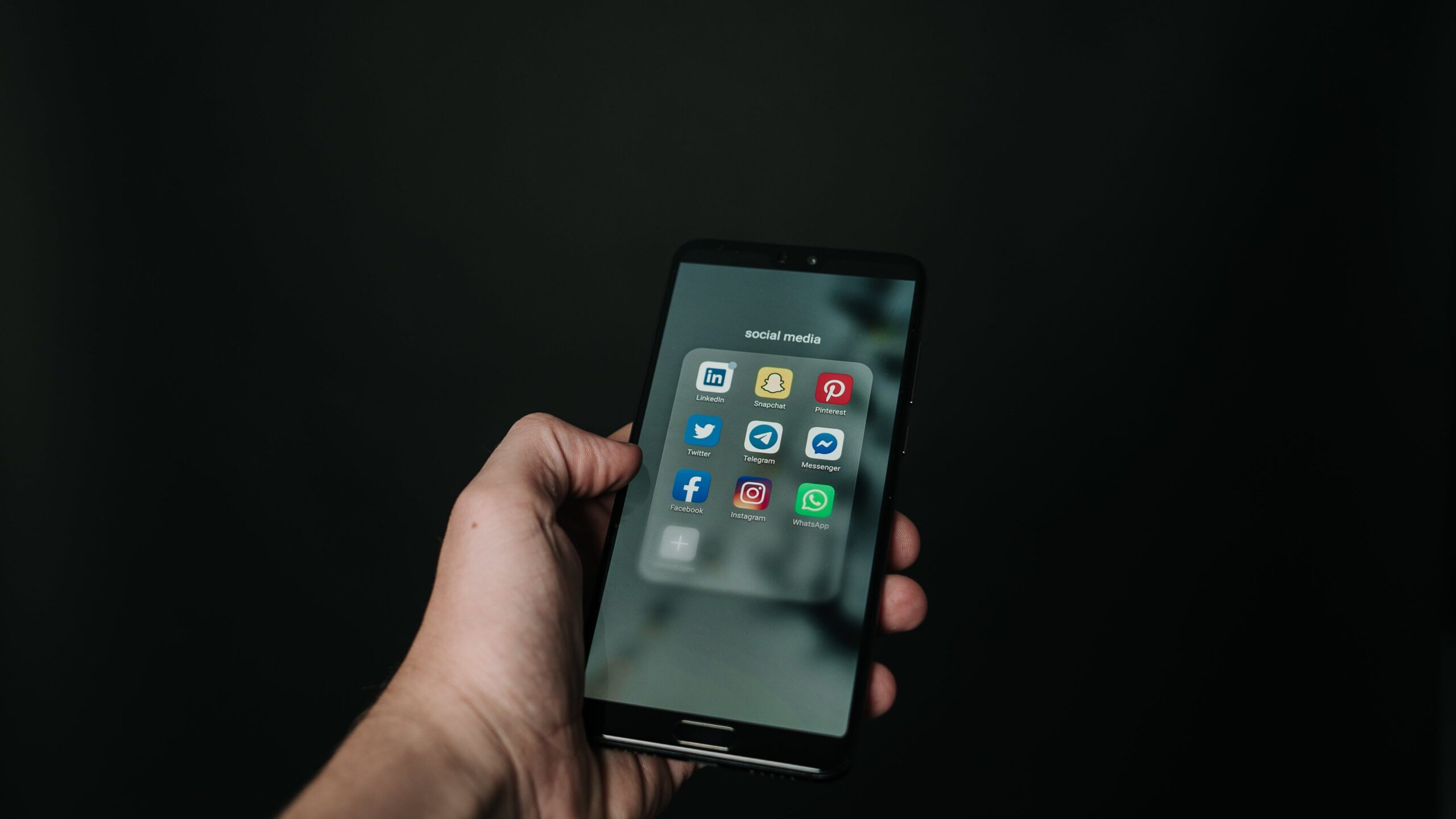 Hand holding smartphone displaying social media apps against a dark backdrop, showcasing the power of connectivity and digital marketing platforms including Twitter, Pinterest, LinkedIn, YouTube, Facebook, Instagram, and WhatsApp, emphasizing the extensive reach of social media marketing for businesses and entrepreneurs.