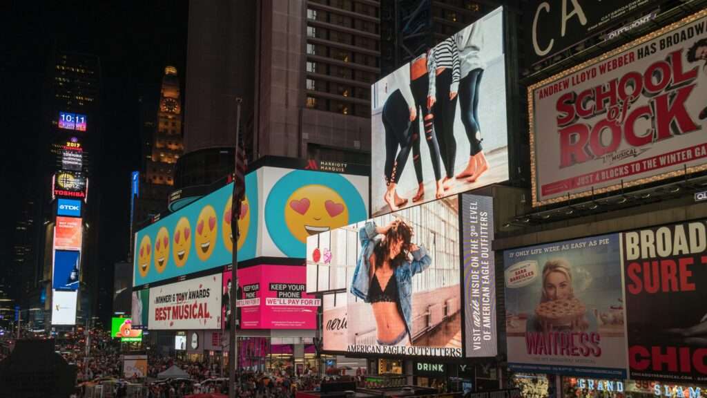 How to Develop an Advertising Strategy: The image depicts a vibrant urban scene with large digital billboards and advertisements at night. It showcases various ads, including emoji faces, a fashion ad with models in leggings, a promotional banner for the musical "School of Rock," and other colorful displays. This bustling area is filled with people and illuminated by the bright lights of the advertisements, highlighting the dynamic nature of modern advertising.